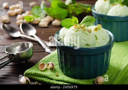 Pistazien-Eiscreme-Kugeln in Tonschüsseln auf einem hölzernen Küchentisch mit Krümel und Nüssen zur Seite Stockfoto