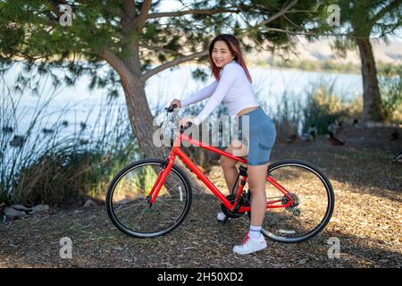 Junge asiatische Frau sitzt auf einem Orange Hybrid Fahrrad vor einem See Stockfoto