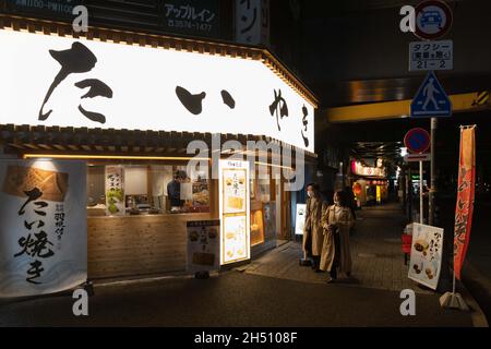 Tokio, Japan. November 2021. Ein Paar, das Gesichtsmasken trägt, als Vorsichtsmaßnahme gegen die Ausbreitung von Covid-19, geht am Takoyaki-Restaurant im Shimbashi-Bezirk vorbei. Kredit: SOPA Images Limited/Alamy Live Nachrichten Stockfoto
