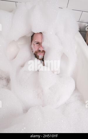 Zufriedener Mann ist fast vollständig mit üppiger Seifenlauge bedeckt, er backt im Bad, genießt und entspannt. Draufsicht. Stockfoto