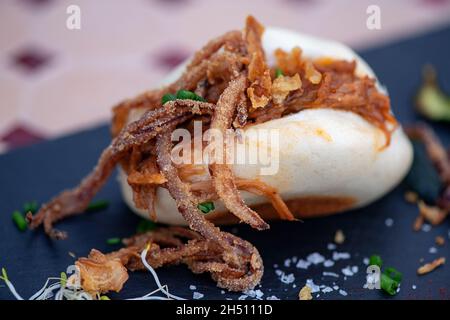 Gedämpftes bao-Brötchen gefüllt mit Schweinefleisch und karamellisierten Zwiebeln und garniert mit Schnittlauch und Meersalz, das auf einem Schieferteller ruht, köstlichen Gourmet-Tapas Stockfoto