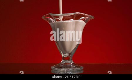 Nahaufnahme von Joghurt in einer Glasschale auf rotem Hintergrund. Archivmaterial. Frischer Joghurt wird in klares Glas gegossen. Stream von Smoothies füllt Glas auf isoliert Stockfoto