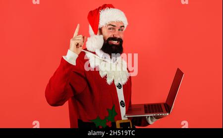 Weihnachtsmann mit Laptop. Bärtiger Geschäftsmann in Santa-Kostüm mit Computer nach oben zeigt. Online-Shopping. Digitales Gerät. Stockfoto