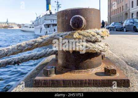 Stockholm, Schweden - 17. April 2021: Anlegeseil um verrosteten Poller mitten in Stockholm und die reguläre Linienfähre im Hintergrund Stockfoto