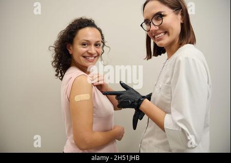 Fröhliche Krankenschwester, medizinische Fachkraft, die auf die Hand der Frau zeigt, mit dem Pfandband, das mit Corona-Virus-Impfstoff geimpft wurde, beide lächelnd und schauend auf t Stockfoto