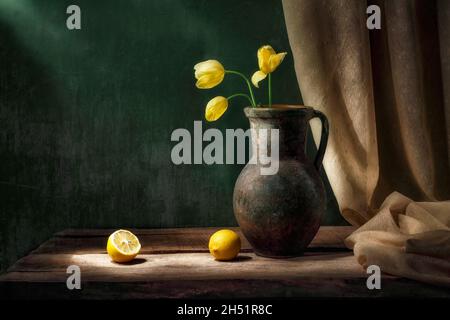 Klassisches Stillleben mit einem Bouquet aus drei gelben Tulpenblüten in einem alten Jahrgangskrug, zwei geschnittenen Zitronenfrüchten und einem Vorrat aus hellem Licht auf grünem Backgroun Stockfoto