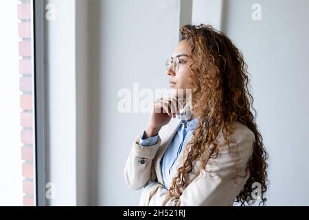 Nachdenkliche Geschäftsfrau aus dem Biracial, die durch das Fenster schaut, während sie im Büro mit der Hand auf dem Kinn steht Stockfoto