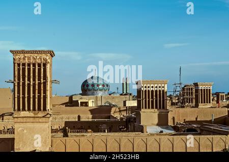 Blick auf die Dächer der alten Stadt Yazd mit Windtürmen -Badgir, mit einer gemalten runden Kuppel einer schönen Moschee und Minarette gegen die BL Stockfoto