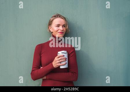 Horizontales Porträt einer attraktiven blonden Frau in den 30ern, die eine wiederverwendbare Tasse Kaffee in der Nähe einer grünen Wand hält Stockfoto
