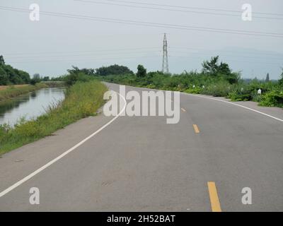 Bewässerungs- und Transportsystem, offene Kurve Asphaltstraße entlang des Kanals mit Berg- und Hochspannungsturm in Thailand Stockfoto