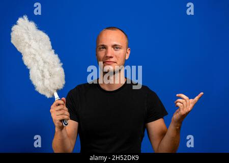 Halblanges Porträt eines jungen lächelnden Hausmeisters in schwarzen T-Shirt-Overalls mit blauem Staubwedel. Isoliert auf blauem Hintergrund Stockfoto