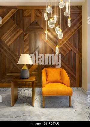 Moderner orangefarbener Sessel, Lampenschirm auf einem kleinen Holztisch und moderner großer Kronleuchter aus Glas in einem Flur mit dekorierter Holzverkleidung und weißem Marmorfußboden Stockfoto