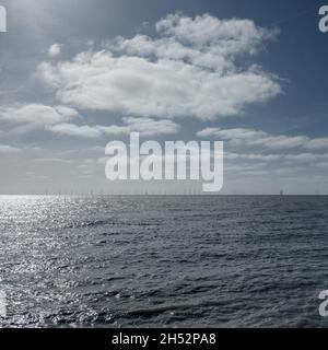 Offshore-Windenergieanlagen am Horizont in Clacton-on-Sea, Essex, mit weißen Wolken und grau-blauem Himmel im Hintergrund und grauem Meer im Vordergrund. Stockfoto