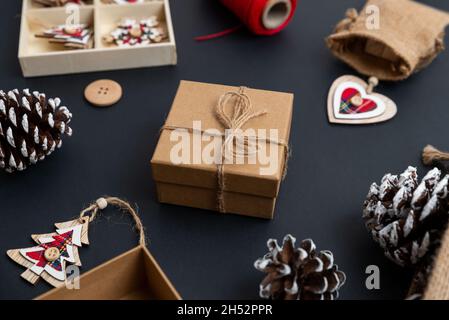 Weihnachtsgeschenk mit Garnschleife auf dem Schreibtisch, umgeben von weihnachtsdekorationen. Tannenzapfen, Holzdekorationen, roter Faden, Sack auf dem Tisch. Nahaufnahme Stockfoto