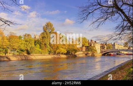 Eine Flussuferszene im Frühherbst. Eine historische Brücke befindet sich neben einem alten Turm und Bäume säumen das Ufer. Ein blauer Himmel mit Wolken ist oben. Stockfoto