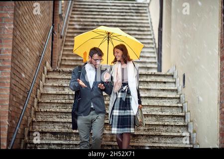 Ein junges verliebes Paar, das während eines regnerischen Tages die Treppe in der Stadt bei guter Laune hinuntersteigt und redet. Gehen, Regen, Stadt, Beziehung Stockfoto