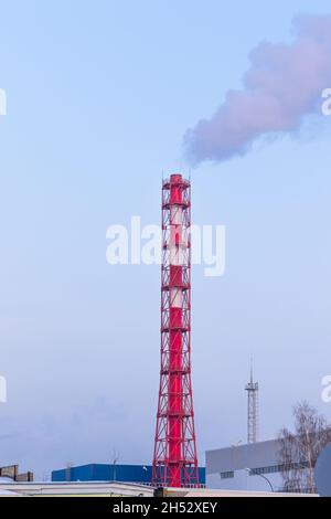 Rot-weißer Schornstein einer Fabrik, aus dem an einem sonnigen frostigen Wintertag grauer Rauch gegen einen blauen Himmel ausläuft Stockfoto