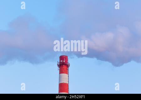 Rot-weißer Schornstein einer Fabrik, aus dem an einem sonnigen frostigen Wintertag grauer Rauch gegen einen blauen Himmel ausläuft Stockfoto