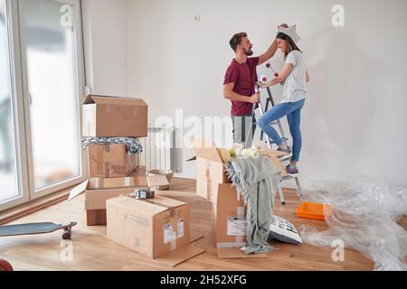Nettes kaukasisches Paar, das Spaß hat, während es sich vorbereitete, weiße Wände in einer neuen Wohnung zu malen, auf einer Leiter stehend, Pinsel haltend, lächelnd, schauend ea Stockfoto