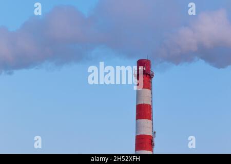 Rot-weißer Schornstein einer Fabrik, aus dem an einem sonnigen frostigen Wintertag grauer Rauch gegen einen blauen Himmel ausläuft Stockfoto