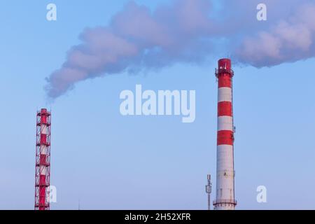 Rot-weißer Schornstein einer Fabrik, aus dem an einem sonnigen frostigen Wintertag grauer Rauch gegen einen blauen Himmel ausläuft Stockfoto