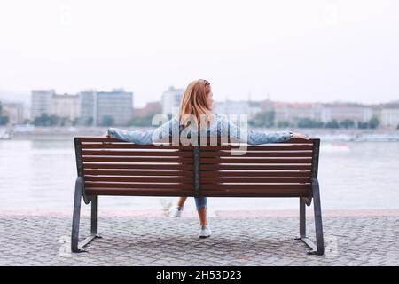 Junge Rothaarige, die sich auf der Bank bei der Rückansicht am Fluss entspannt Stockfoto