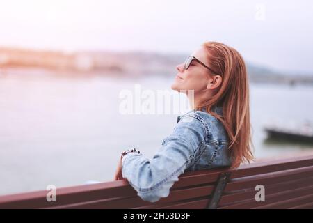 Junge, ausgeglichene, rothaarige kaukasische Frau, die auf einer Bank am Flussufer sitzt und bei Sonnenuntergang meditiert Stockfoto