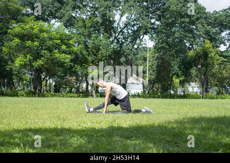 Sportlich aktiver junger Mann in Sportbekleidung, der im Park Sportübungen macht. Passen kaukasischen Mann Stretching auf der schwarzen Sportmatte im Freien. Stockfoto