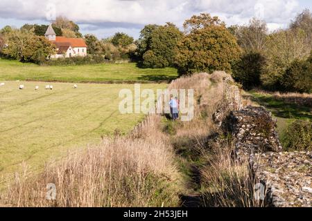 Touristen, die neben einem Teil der ruinierten römischen Stadtmauer in Silchester mit der Kirche der Jungfrau Maria im Hintergrund spazieren. Hampshire, Großbritannien Stockfoto