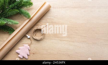 Weihnachtliches rosafarbenes Spielzeug in Form eines Baumes, einer Rolle Bastelpapier und einem Tannenzweig auf einem hölzernen Hintergrund mit Kopierfläche, flach liegend. Umweltfreundliche Verpackung Stockfoto