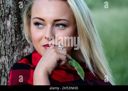 Die junge blonde Frau schaut nach links, während sie sich an einem Baum lehnt und sich mit einem grünen Blatt im Freien streichelt Stockfoto