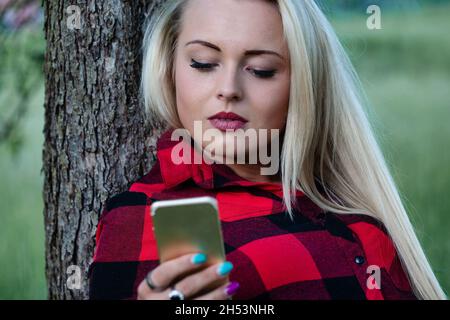 Wunderschöne blonde Mädchen lehnt sich an einen Baum überprüft ihren Handy-Status, weil sie nicht wollen, Anhänger zu verlieren Stockfoto