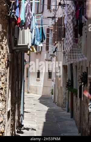 Koper, Slowenien - 12. Juli 2021: Enge Straße zwischen alten, baufälligen Stadthäusern. Kleidung hing zwischen Gebäuden. Stockfoto