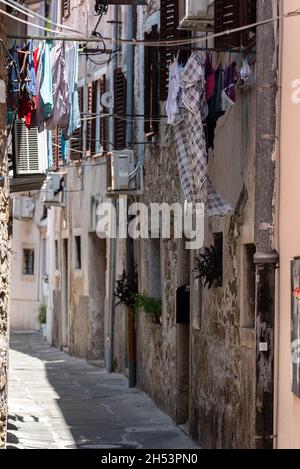 Koper, Slowenien - 12. Juli 2021: Enge Straße zwischen alten, baufälligen Stadthäusern. Kleidung hing zwischen Gebäuden. Stockfoto