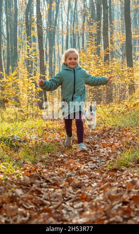 Fröhliches Kind Mädchen in Jacke läuft vom Hund auf dem Trail im Herbstwald. Unter den Füßen des Kindes sind Blätter gefallen. Baumstämme im Hintergrund Stockfoto