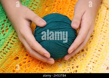 Blauer Wollball in den Händen vor dem Hintergrund einer grün-gelben gestrickten Tischdecke. Hochwertige Fotos Stockfoto