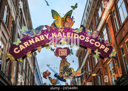 Carnaby Street, London, Großbritannien. November 2021. Die Carnaby Street ist voll von Käufern und Besuchern, die die diesjährige Installation, das Carnaby Kaleidoskop, bewundern, die diese Woche enthüllt wurde. Über 600 handverzierte funkelnde Schmetterlinge mit Neonmustern flattern an Weihnachten durch die Carnaby Street. Der Carnaby-Bogen leuchtet hell in rosa Schimmerscheiben und ein fröhlicher Regenbogen feuert die Besucher an. Carnabys Weihnachtsbeleuchtung in diesem Jahr wird in Zusammenarbeit mit der Wohltätigkeitsorganisation Choose Love durchgeführt, um Flüchtlinge auf der ganzen Welt hervorzuheben und zu unterstützen. Kredit: Imageplotter/Alamy Live Nachrichten Stockfoto