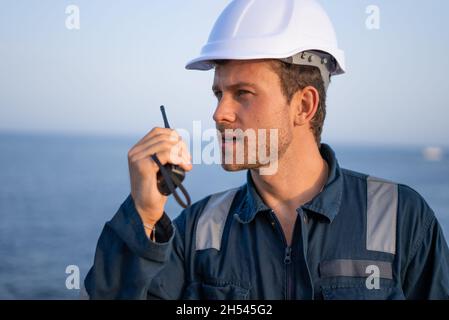 Seriöse junge bärtige männliche Spezialistin für Uniform und schützenden Hardhat, die mit Walkie Talkie spricht, während sie im Seehafen arbeitet Stockfoto