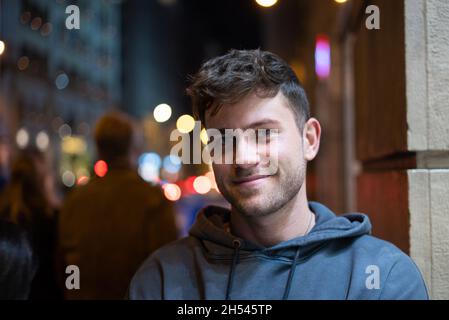 Positiver Mann im lässigen Hoodie, der lächelt und auf die Kamera schaut, während er auf dem unscharfen Hintergrund der Nachtstadt steht Stockfoto
