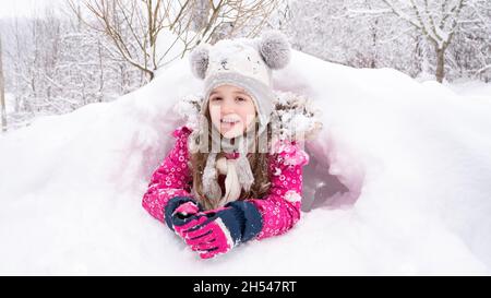 Das Mädchen liegt in einer Schneeverwehung und lächelt. Das Kind in einem lustigen Hut hat ein Iglu oder eine Schneehöhle gebaut. Ideen für Winteraktivitäten für Kinder. Wofür man sich etwas tun kann Stockfoto