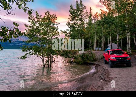 Der perfekte Campingplatz am Abraham Lake mit Bäumen, Zelt und Lastwagen vor dem Sonnenuntergang beleuchteten Himmel mit Bergen im Hintergrund Stockfoto