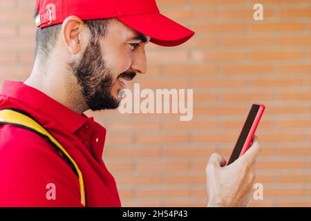 Glücklicher hispanischer Lieferer in roter Kleidung mit Mobiltelefon Stockfoto