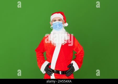 Ein kaukasischer Mann, der als Weihnachtsmann gekleidet ist, trägt eine blaue OP-Maske. Der Hintergrund ist grün. Stockfoto