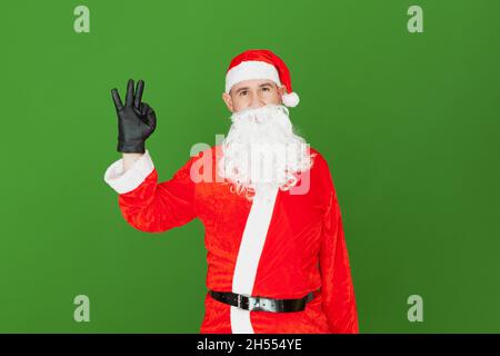 Ein kaukasischer Mann, der als Weihnachtsmann gekleidet ist, macht mit den Fingern seiner Hand eine Daumen-nach-oben-Geste, während er geradeaus blickt. Der Hintergrund ist gree Stockfoto