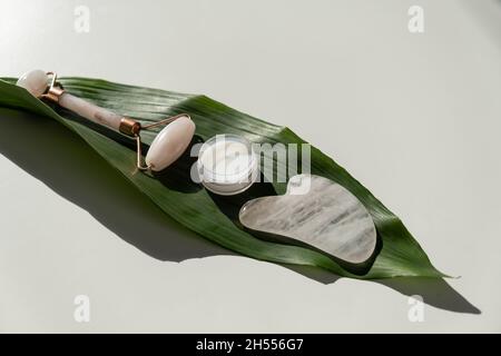 Rosafarbener Gouache-Abstreifer, Walze, Glas mit grüner Blattcreme auf einem weißen Tisch. Ausrüstung für die Selbstmassage und die Pflege von Gesicht und Hals Stockfoto