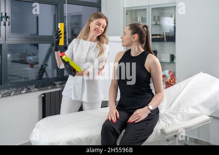Der Kosmetik-Hautarzt hält ein Paket mit Medikamenten zur Gesichtsverjüngung bereit. Eine Ärztin in seinem Büro zeigt den Kunden Behandlungsmöglichkeiten Stockfoto