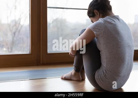Rückansicht junge unglückliche Frau emb Knie sitzen in der Nähe des Fensters Stockfoto