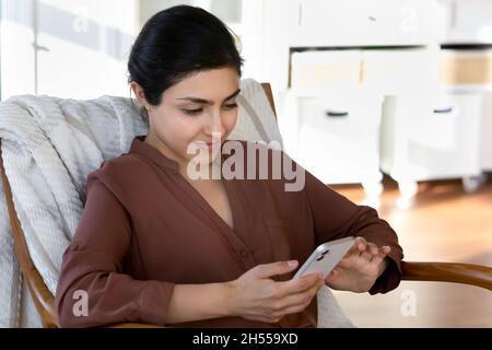 Ruhige schöne indische Frau, die sich auf einem Sessel mit Smartphone drinnen entspannt Stockfoto