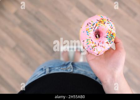 Eine Frau steht auf der Waage, hält einen Donut in den Händen, Draufsicht, Kopierraum. Stockfoto