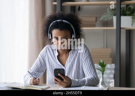 Fokussierte afroamerikanische Frau mit Kopfhörern, die Notizen macht und das Smartphone hält Stockfoto